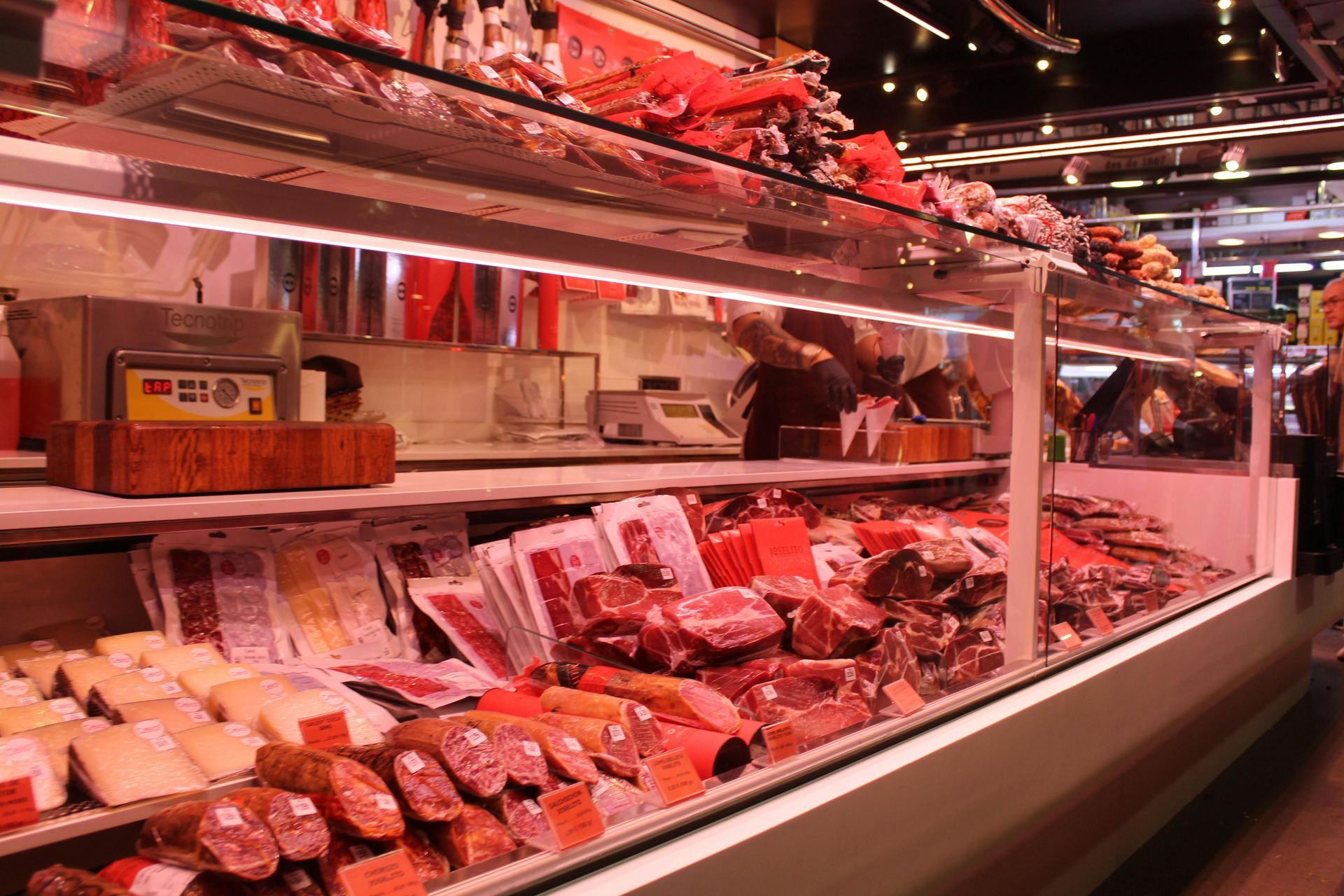 Display case filled with various meats and cheeses in a butcher shop, illuminated by red lighting.