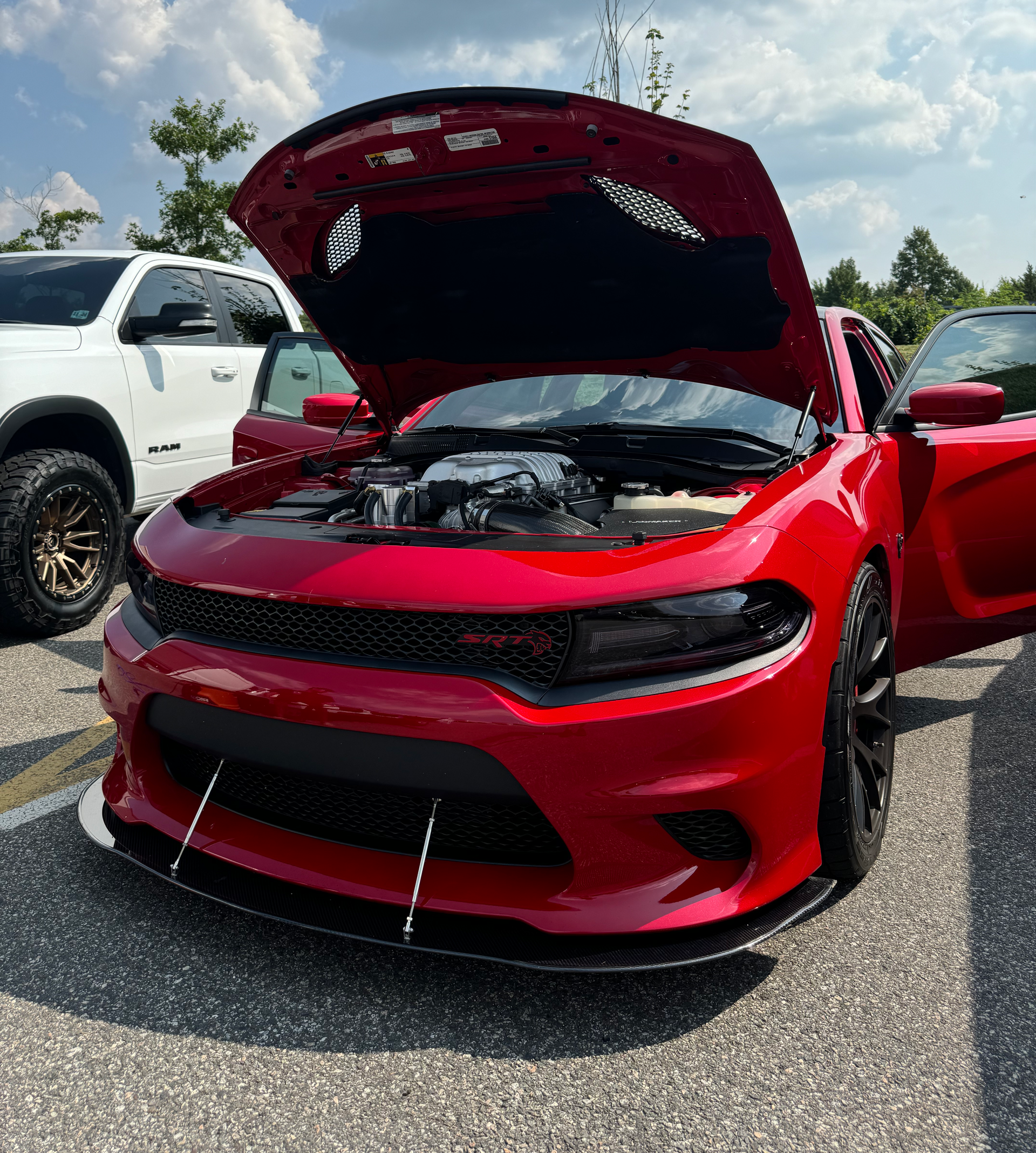 A red Hellcat with the hood up is parked next to a white truck.