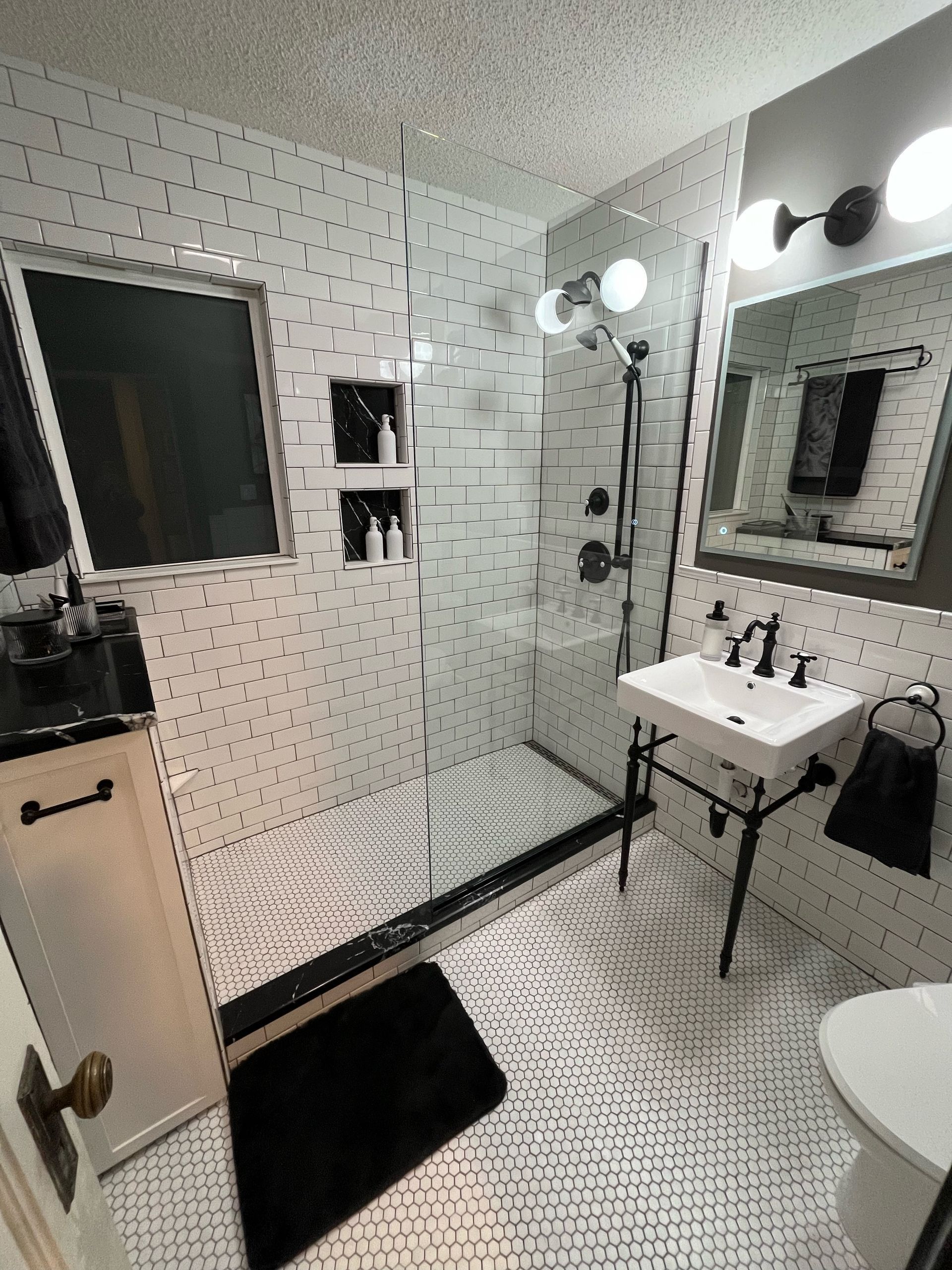 Modern white bathroom with walk-in shower, vanity sink, mirror, and black floor mats.