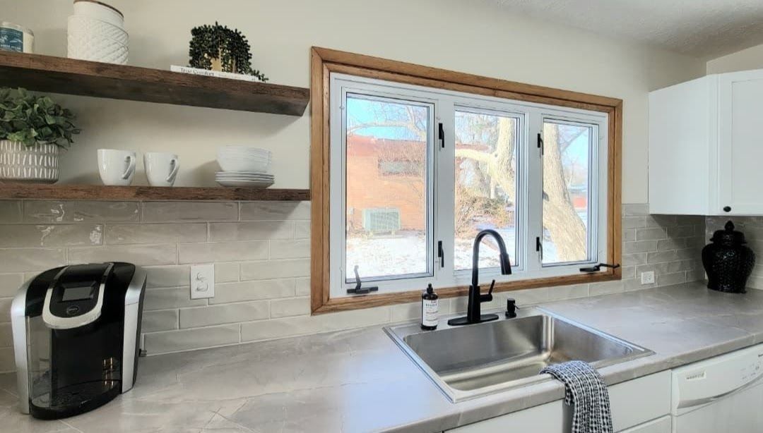 Bright kitchen sink beneath window, white countertops, open shelves, and black coffee maker