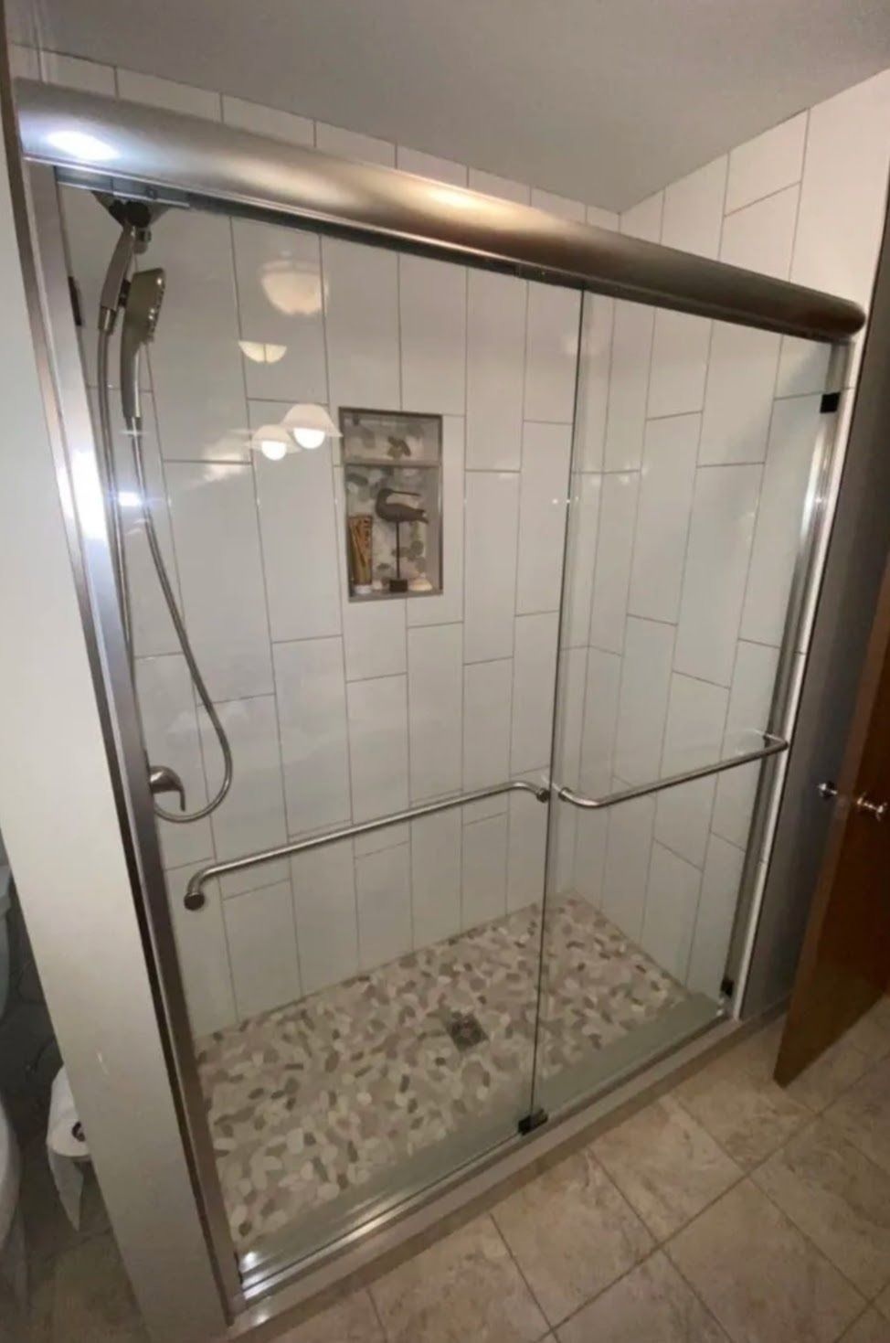 Walk-in shower with glass door, white tiled walls, chrome grab bars, and pebble-style floor.