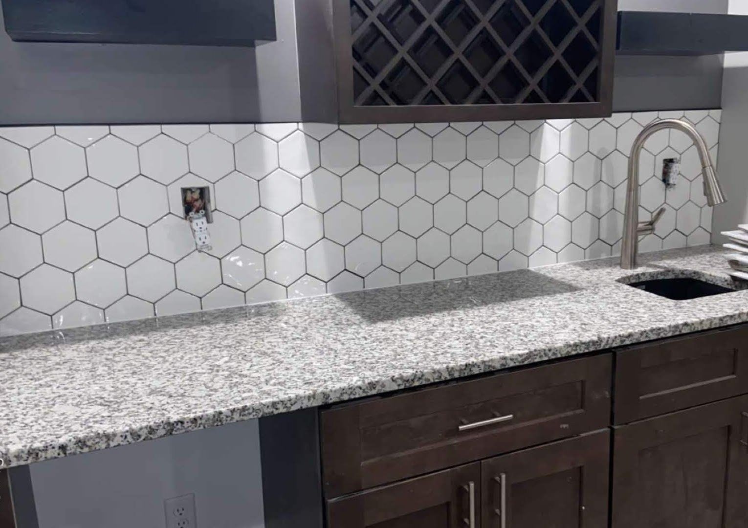 Kitchen countertop with white hex tile backsplash, granite counter, dark cabinets, and stainless sink under a window.
