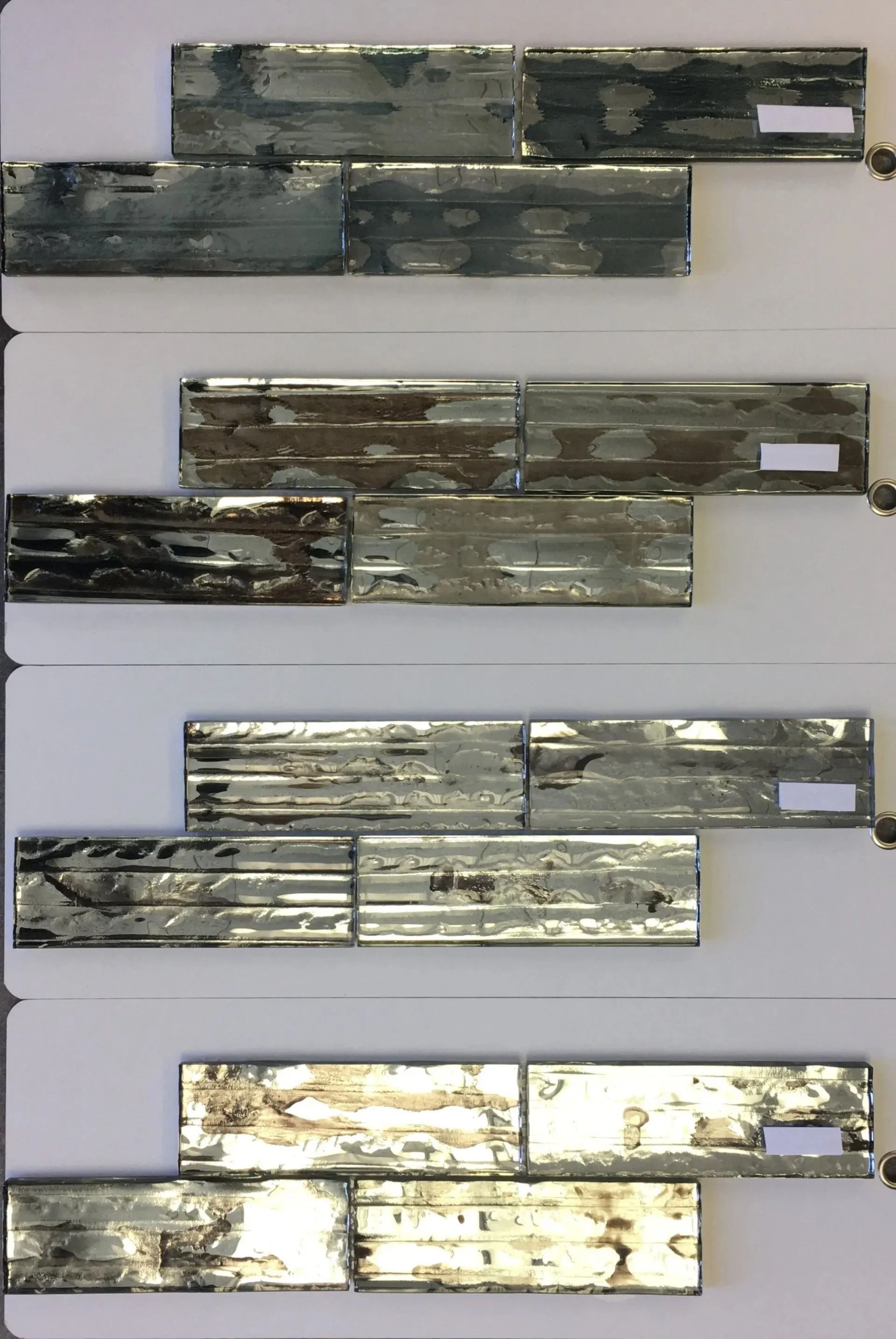 Four stacked samples of metallic tile strips in shades of gray, silver, and gold on a plain background