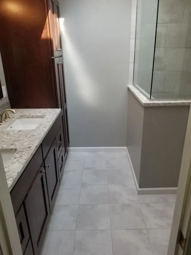 Narrow bathroom with white tile floor, granite countertop, dark wood cabinets, and glass shower enclosure