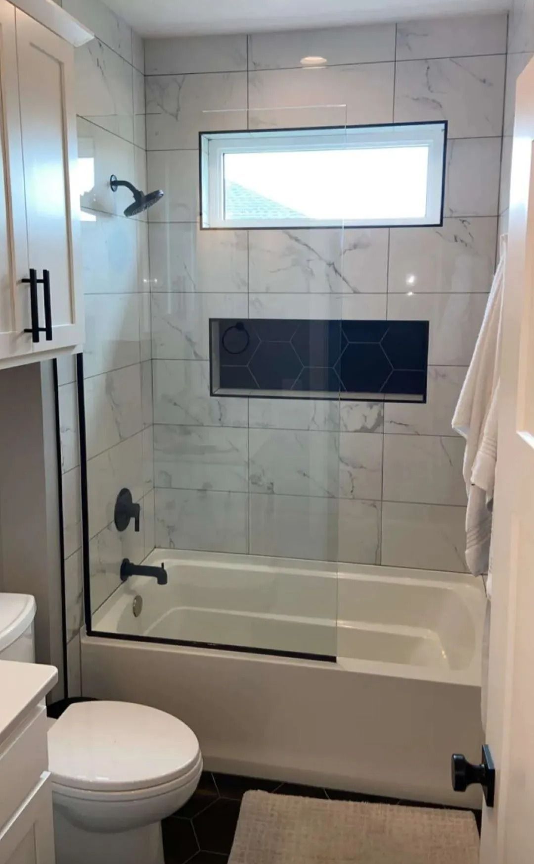 Small tiled bathroom with a tub-shower, frosted window, black fixtures, and a white towel.