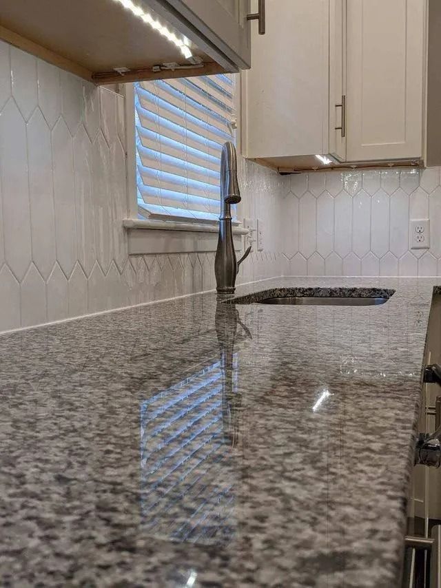 Empty kitchen with granite countertop, white cabinets, sink, and window with blinds.