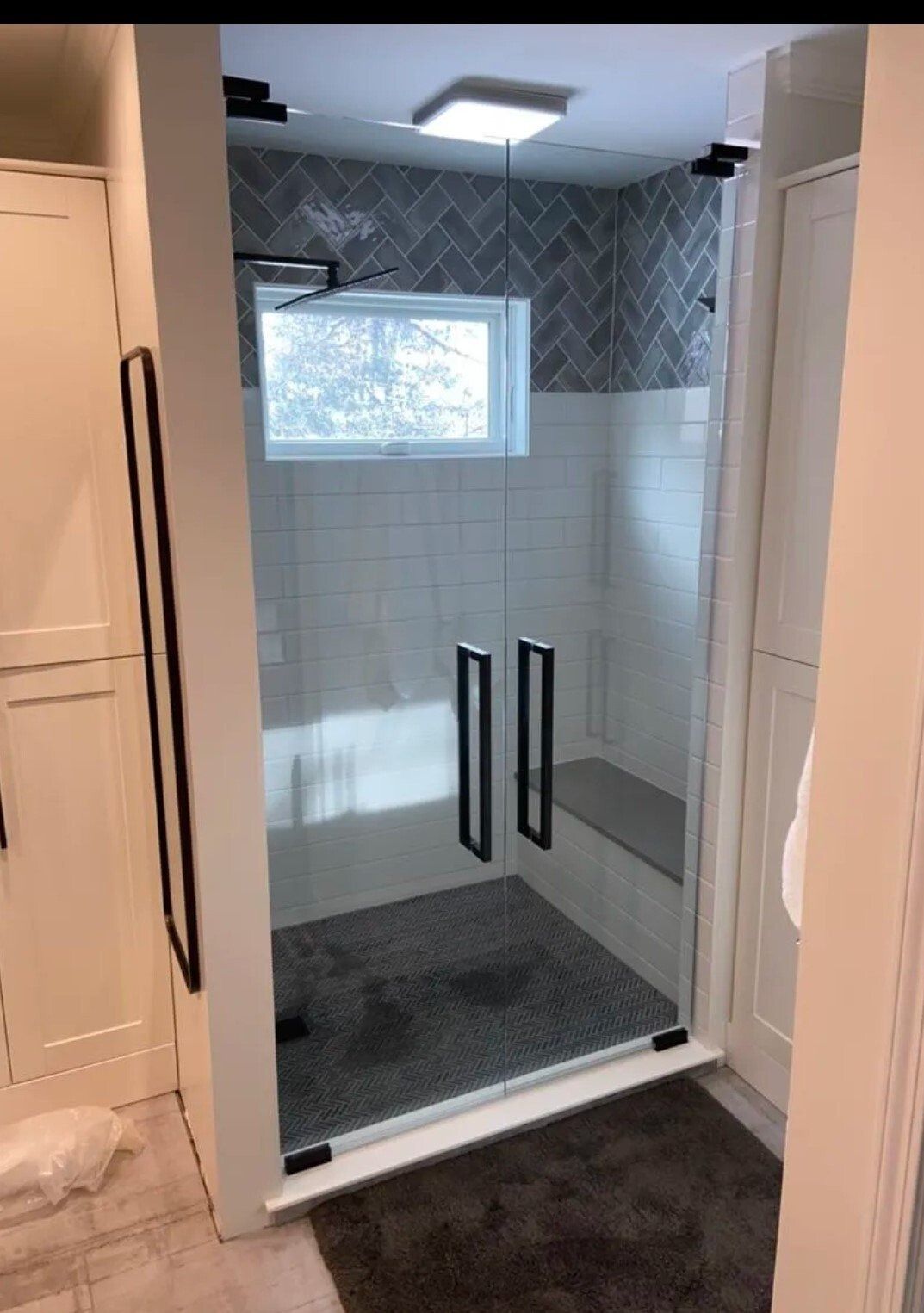 Glass shower enclosure with black handles in a tiled bathroom, lit by a small window.