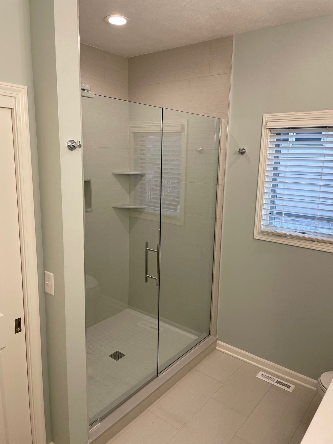 Modern bathroom with glass walk-in shower, beige tile, and a small window with blinds