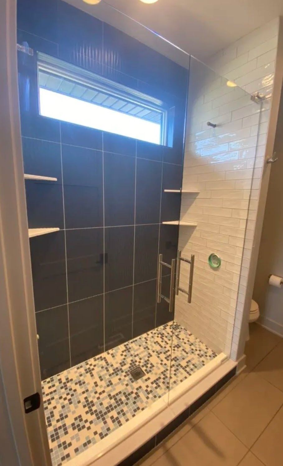 Modern tiled walk-in shower with black wall tiles, mosaic floor, glass door, and built-in shelves