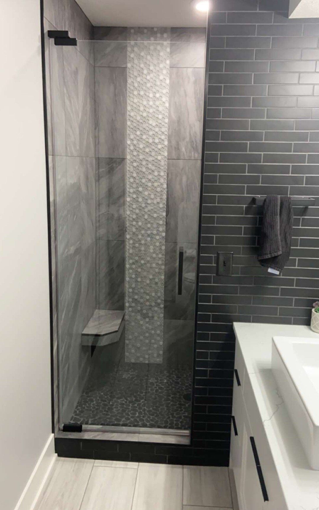 Glass shower stall with gray tile accent wall and black brick wall beside a white vanity sink.
