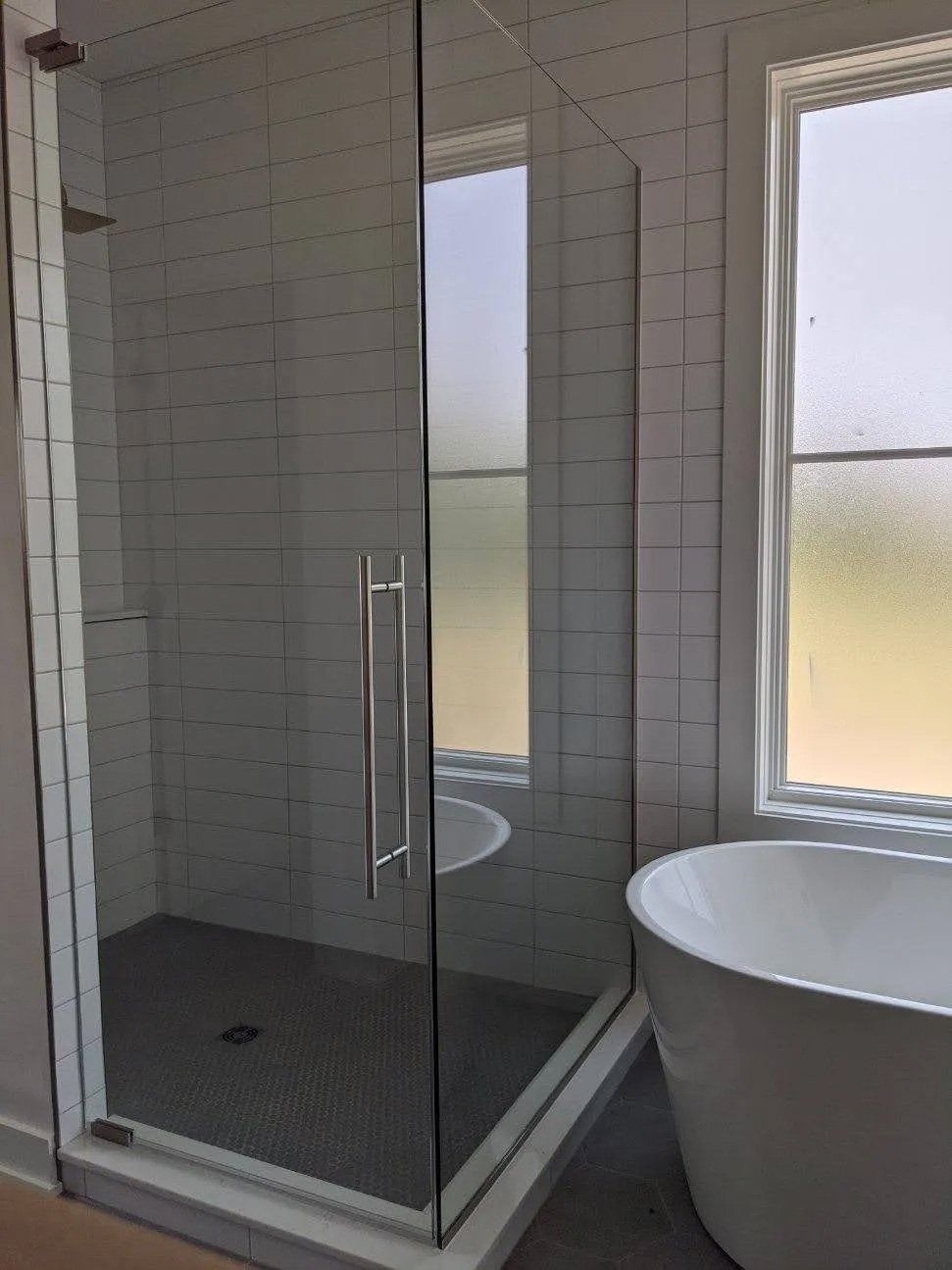 Glass shower enclosure in a white tiled bathroom beside a freestanding tub and frosted window.
