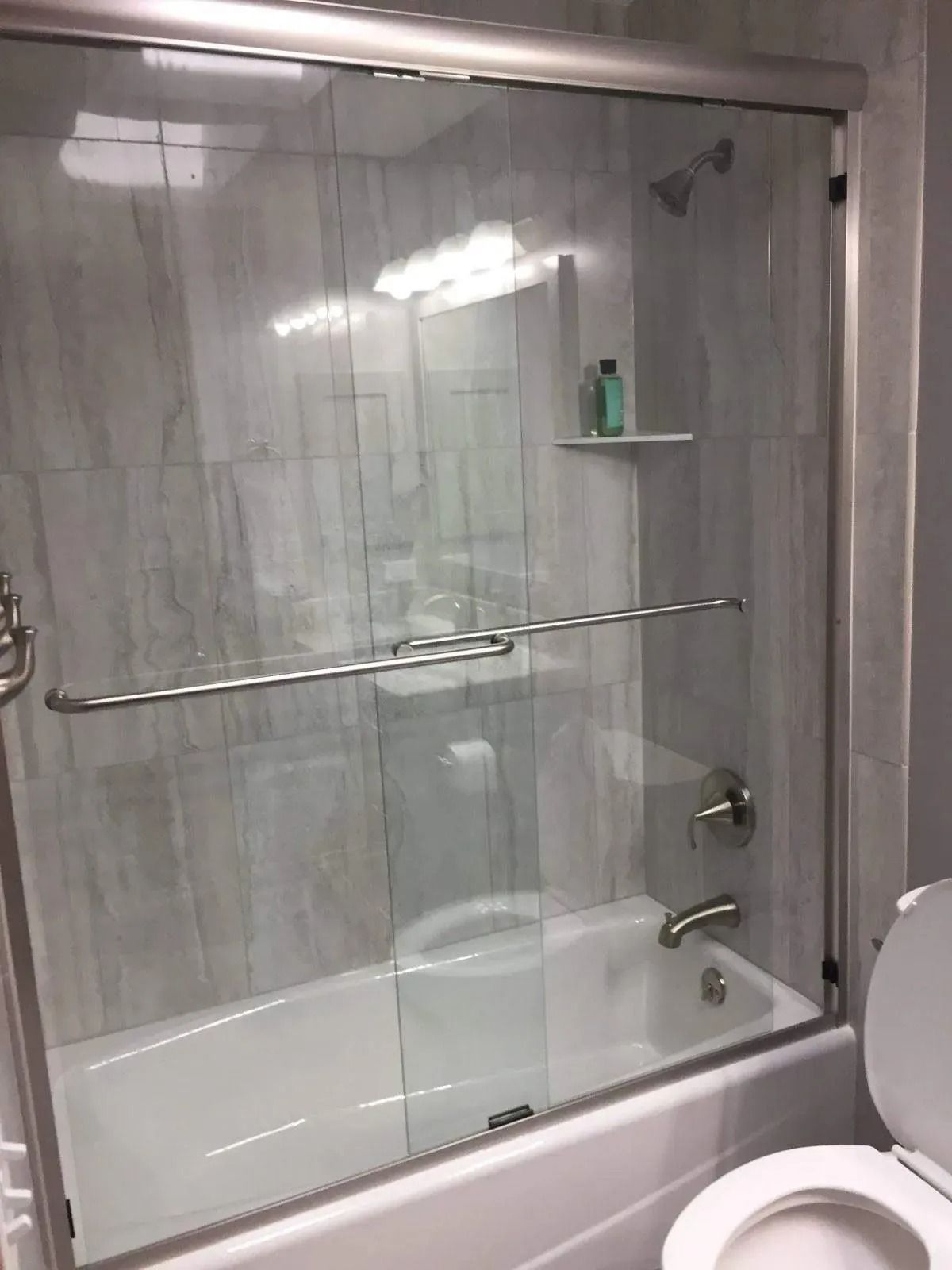 Tiled bathroom with a glass shower-tub enclosure, chrome bar, and a soap shelf.