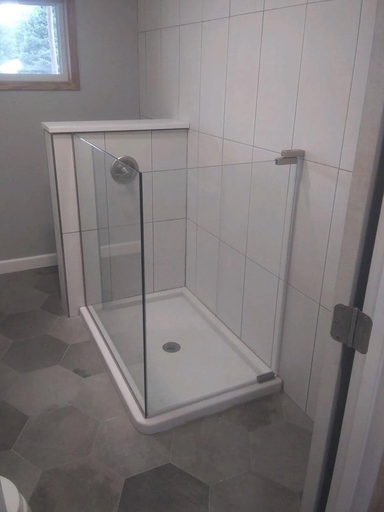 Glass shower stall with white tiled walls and gray hexagonal bathroom floor.