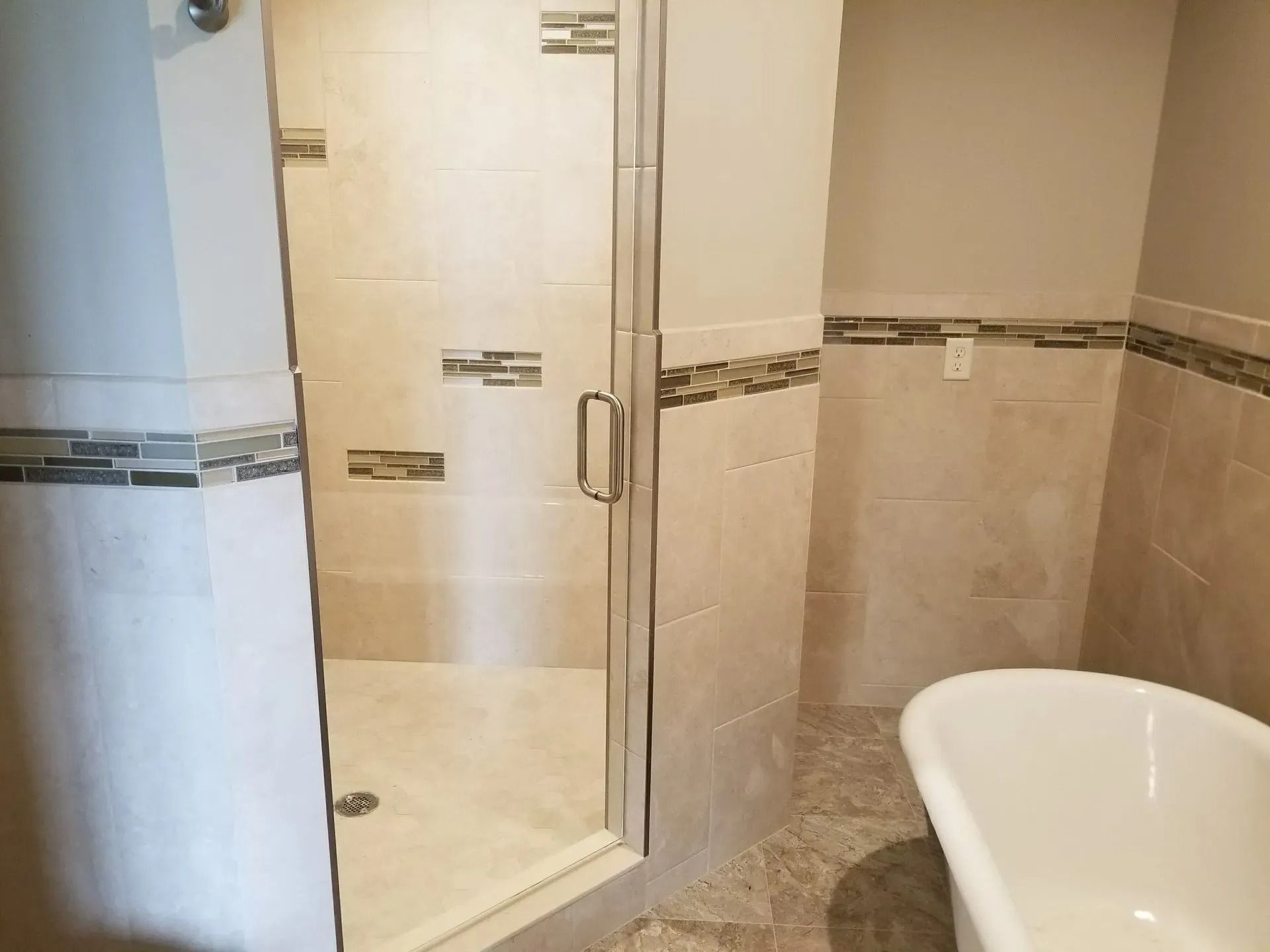 Bathroom with glass shower door, beige tile walls, and white bathtub.