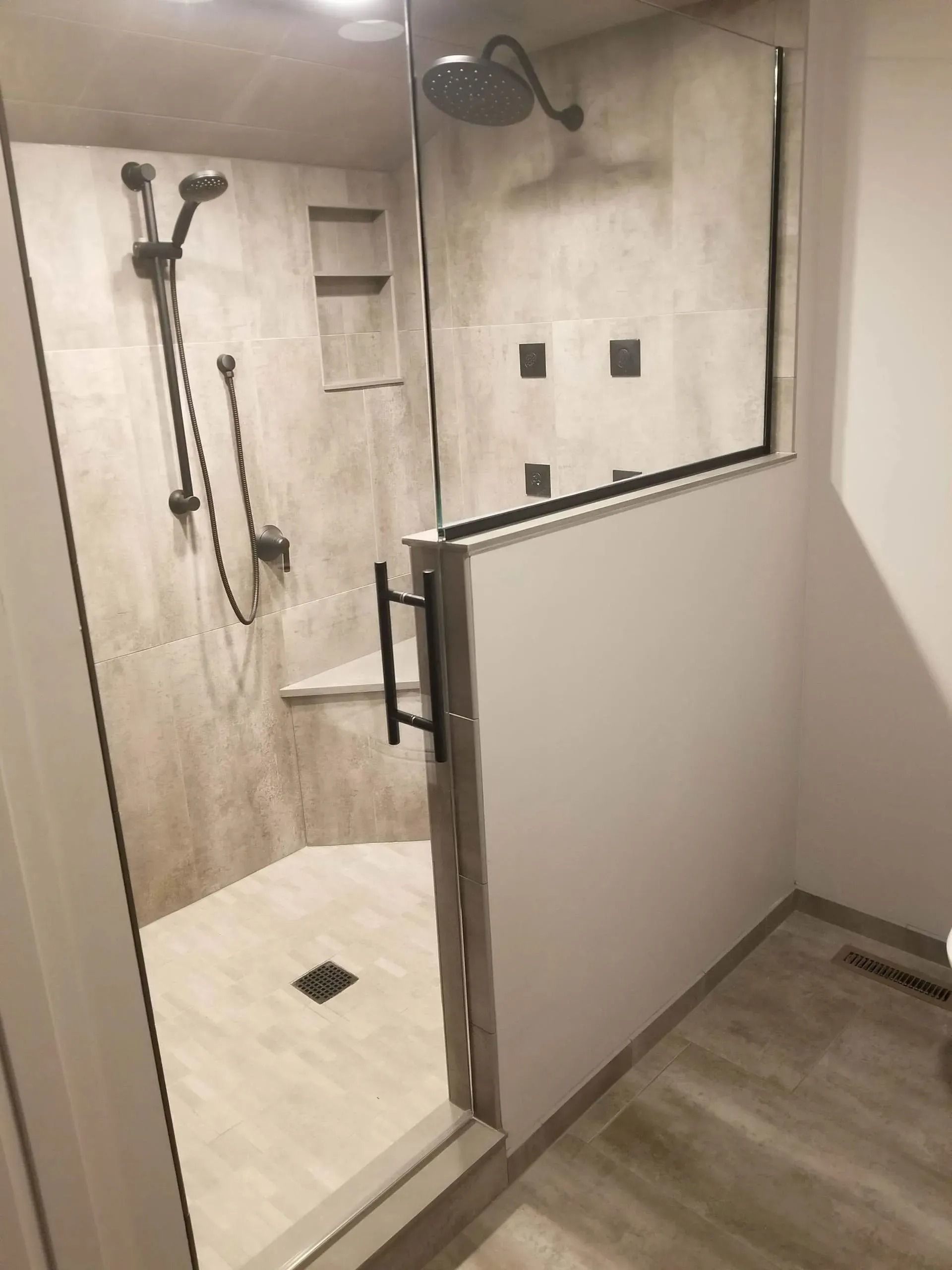 Modern shower with glass partition, beige tile walls, shower fixtures, and a small wall shelf.