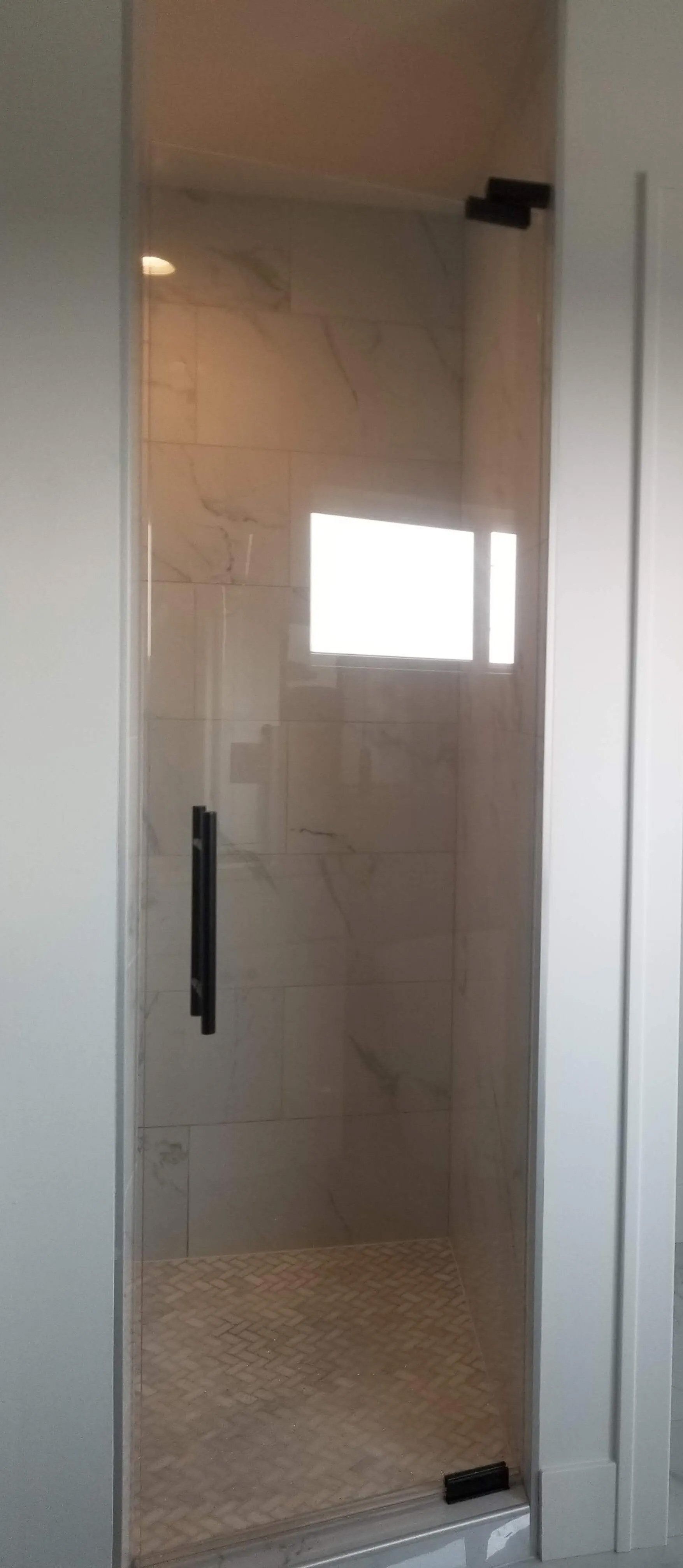 Glass shower door with black handle in a tiled bathroom, showing a bright window reflection