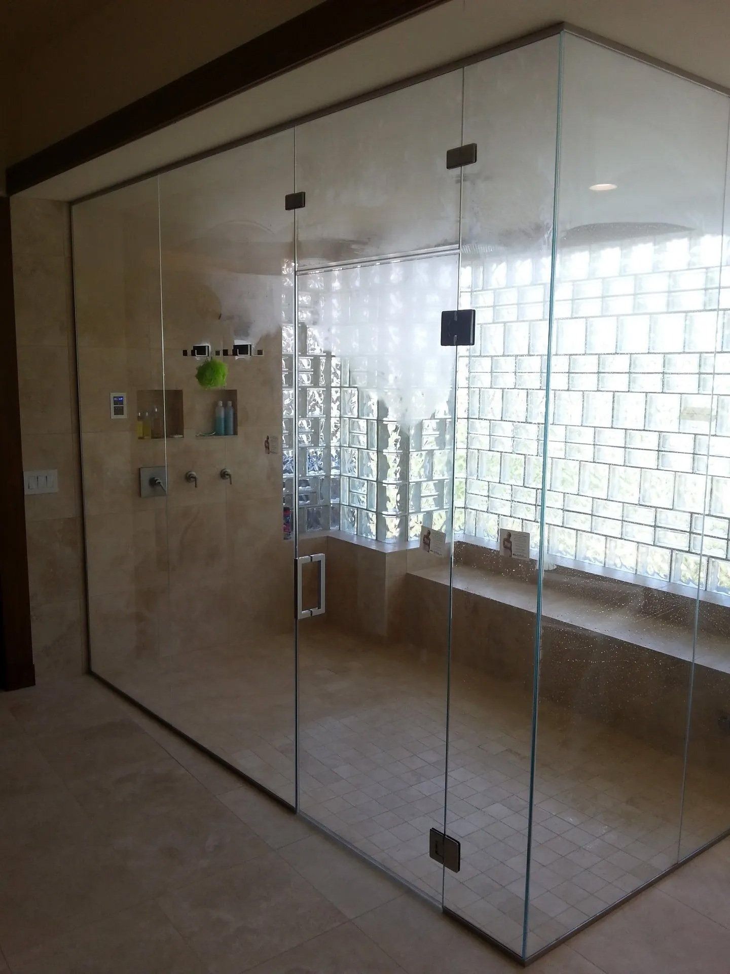 Glass-fronted enclosed shower in a tiled bathroom with a window and wooden bench.