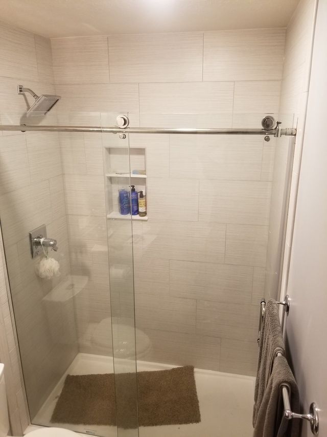 Glass shower stall with beige tiled walls, chrome fixtures, and a brown bath mat