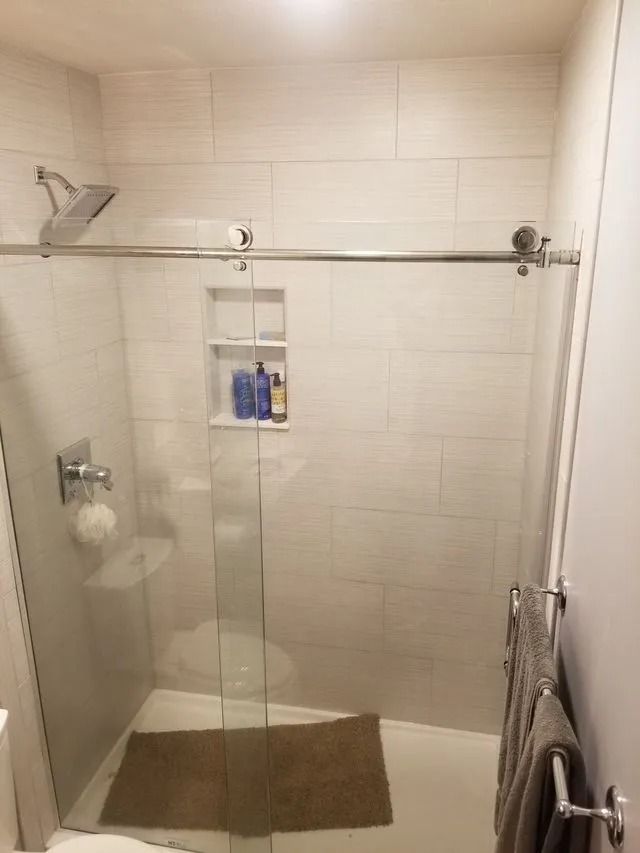 Glass walk-in shower with beige tile, showerhead, grab bar, and wall shelf with toiletries.