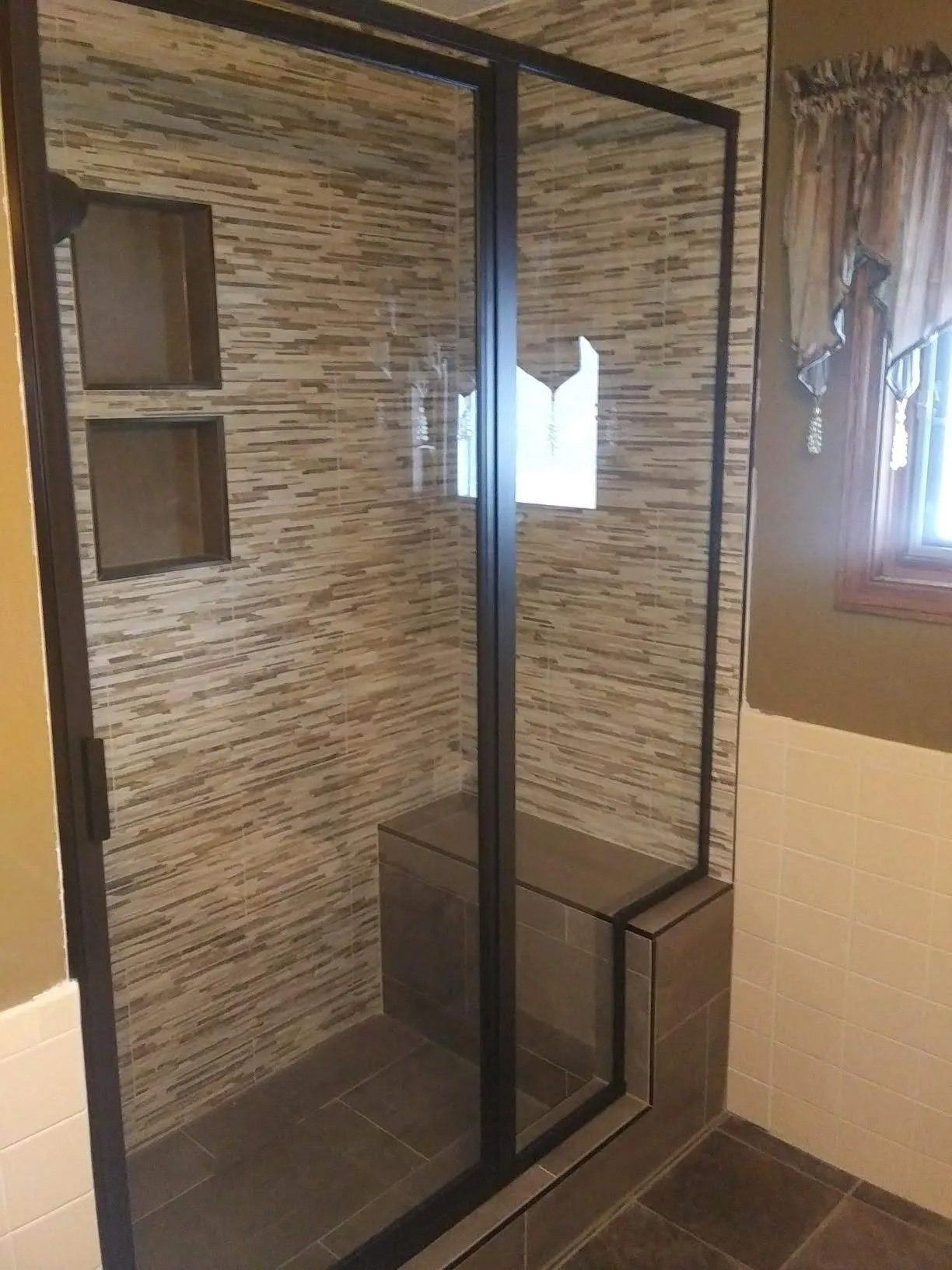 Glass shower enclosure with black frame, gray tile walls, and a small window in a bathroom