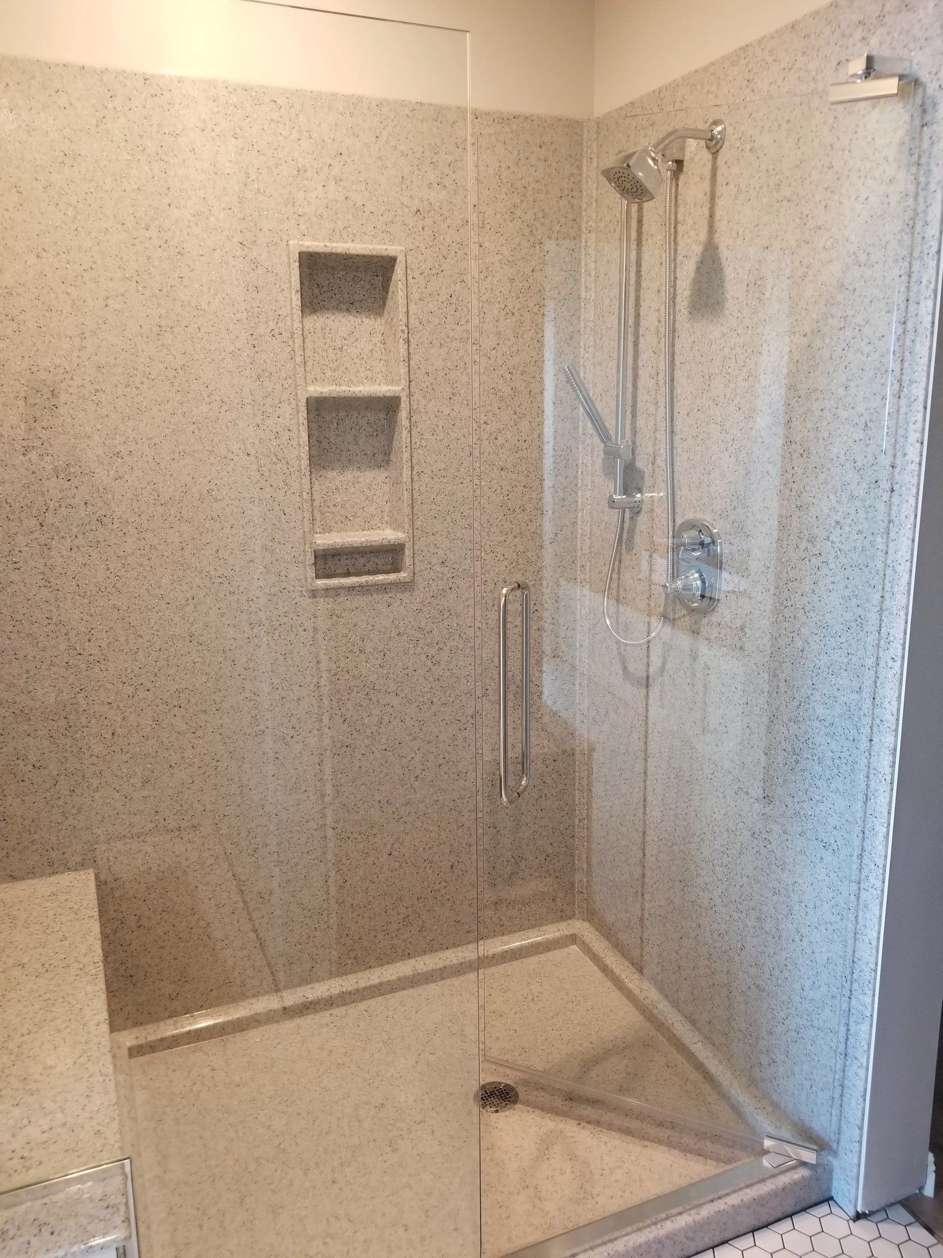 Walk-in shower with speckled tile walls, built-in niches, and glass door partially open.