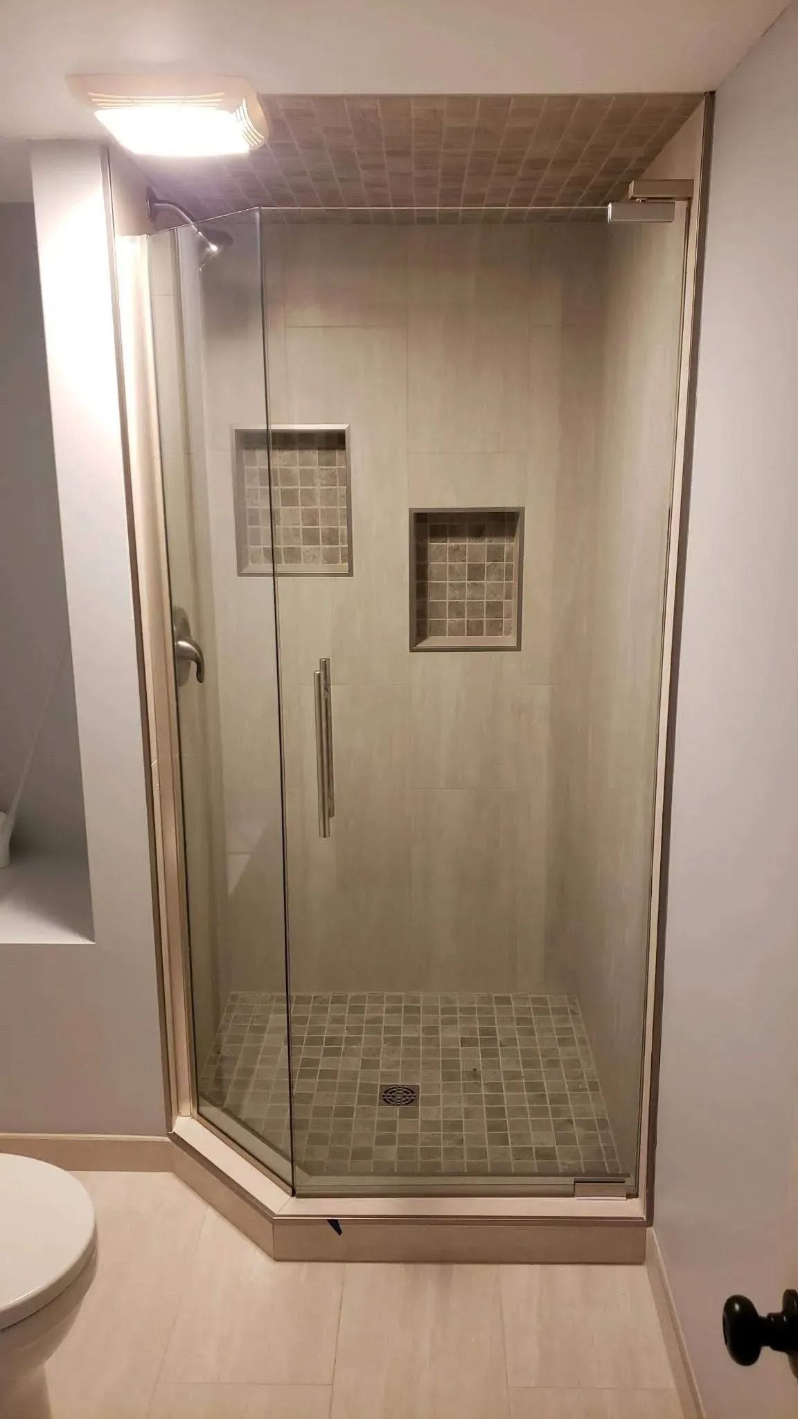 Glass-enclosed shower stall with beige tile walls, mosaic floor, and two small built-in shelves.