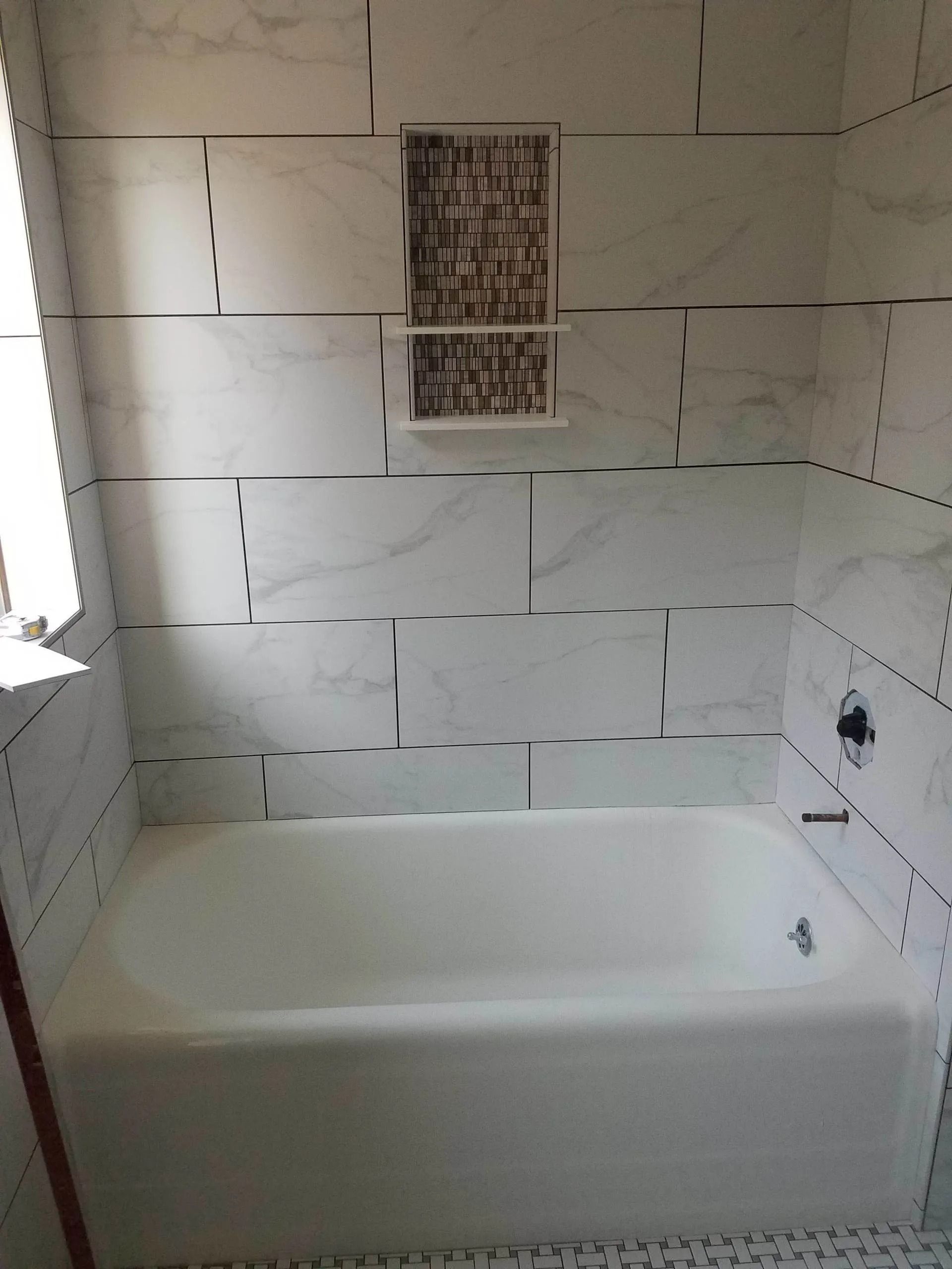 Bathroom tub surrounded by white tiled walls with a decorative accent strip and built-in niche.