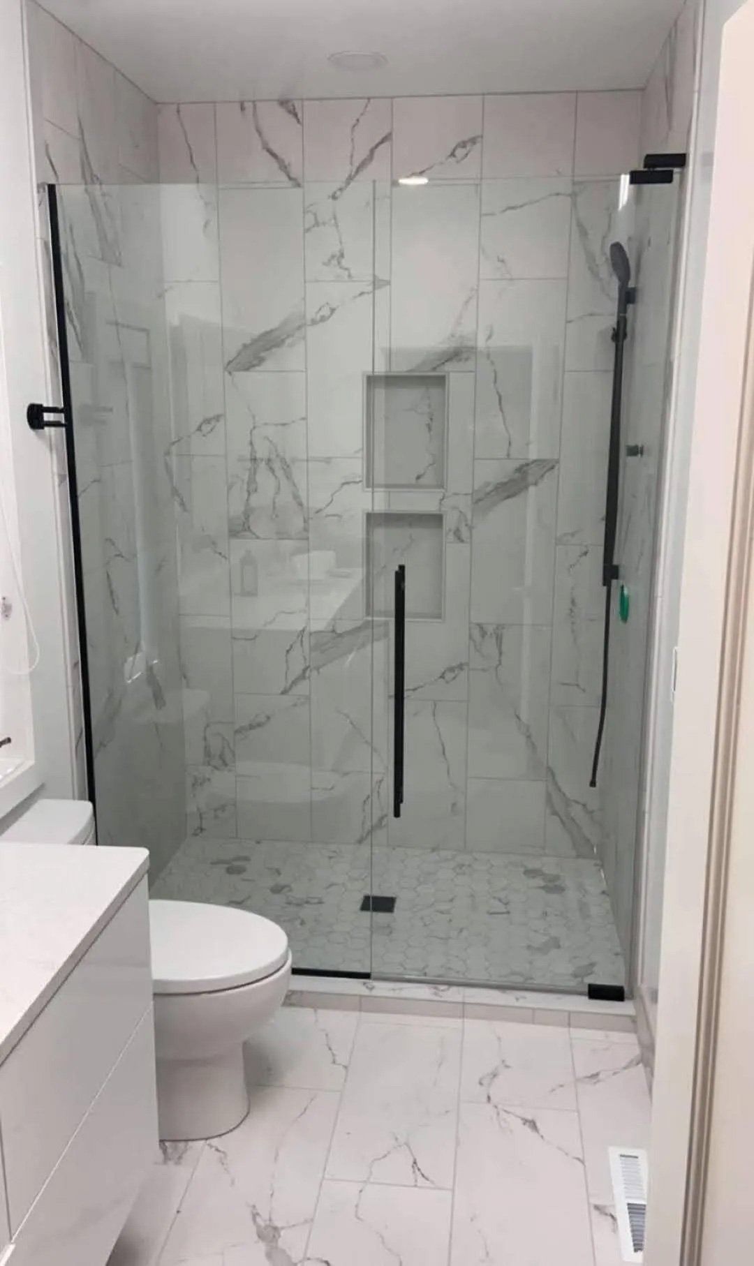 Modern white bathroom with a glass shower enclosure and marble-pattern tile walls and floor.