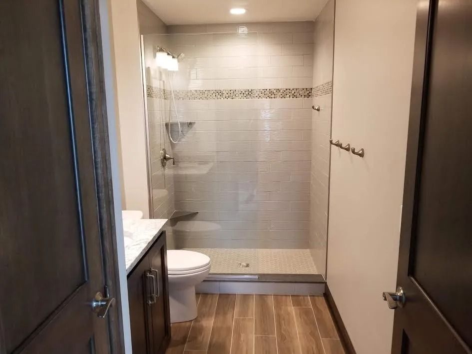 Small bathroom with shower, toilet, sink, and wood floor, viewed through an open door.