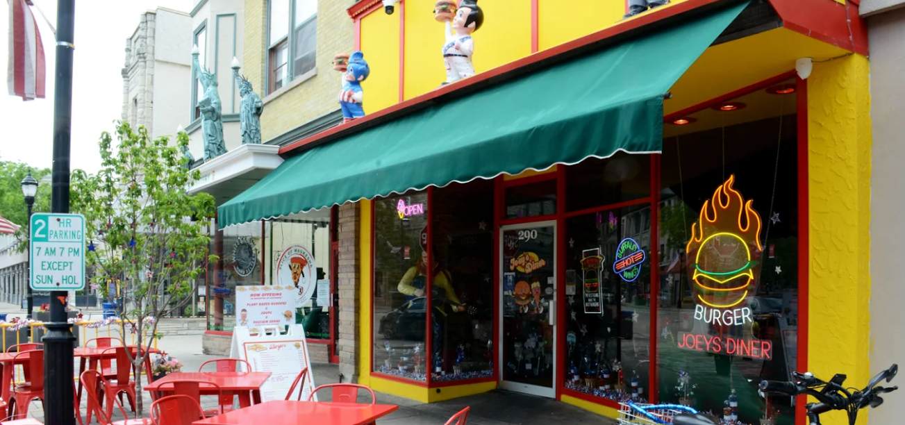 Exterior of a burger restaurant with red tables. Green awning, yellow building, neon burger sign.