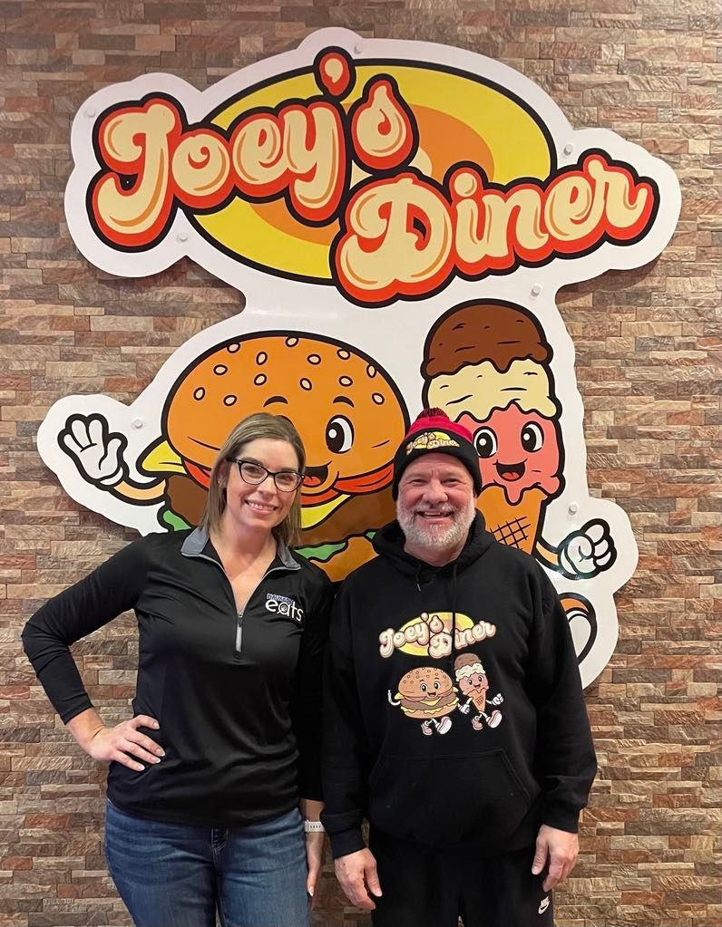 Woman and man stand in front of Joey's Diner sign with burger and ice cream cone graphics.