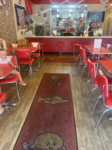 Interior of a diner: Red tables and chairs, long mat, people behind the counter, a child seated at a table.