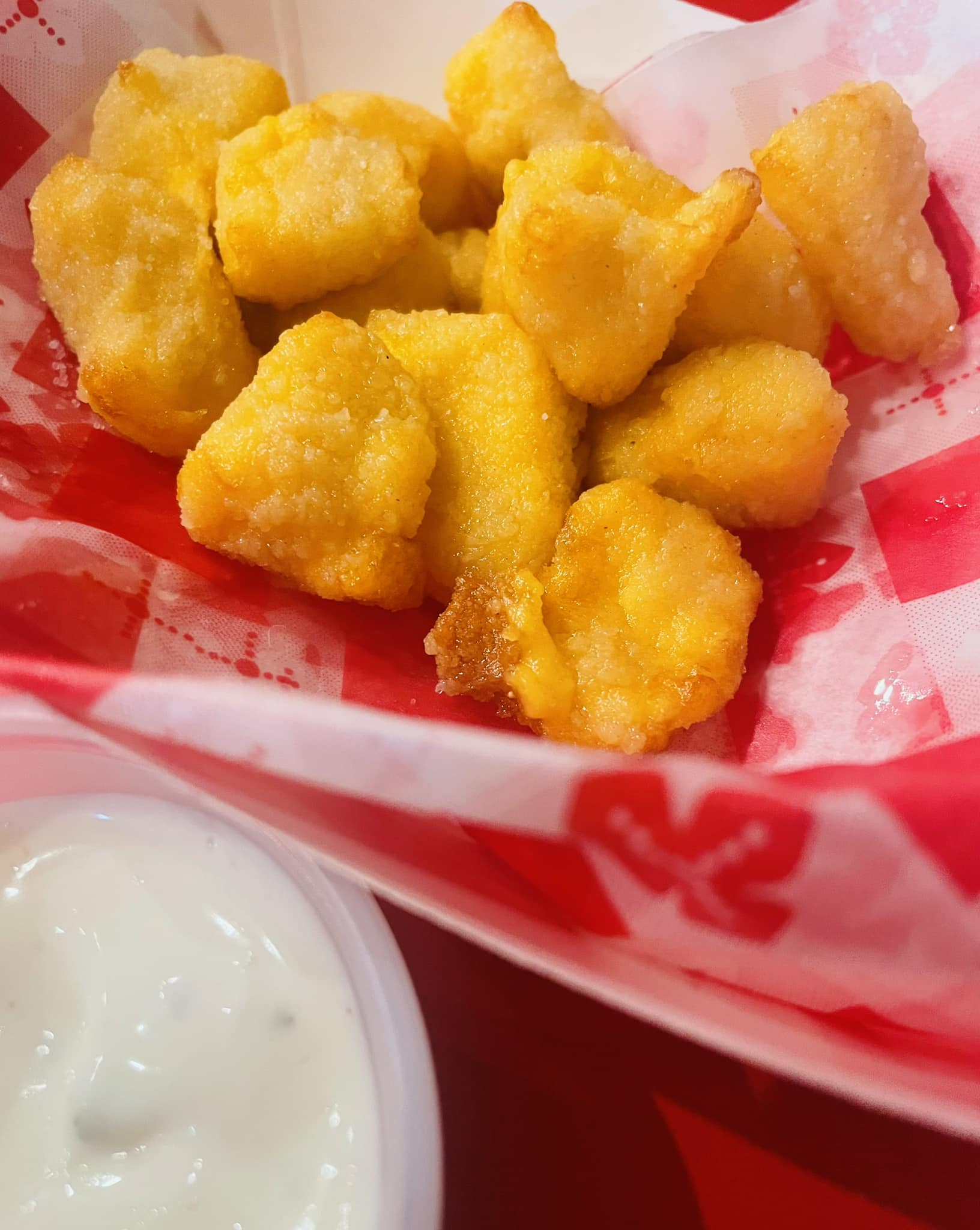 Golden fried cheese curds in a red and white basket, next to a container of white dipping sauce.