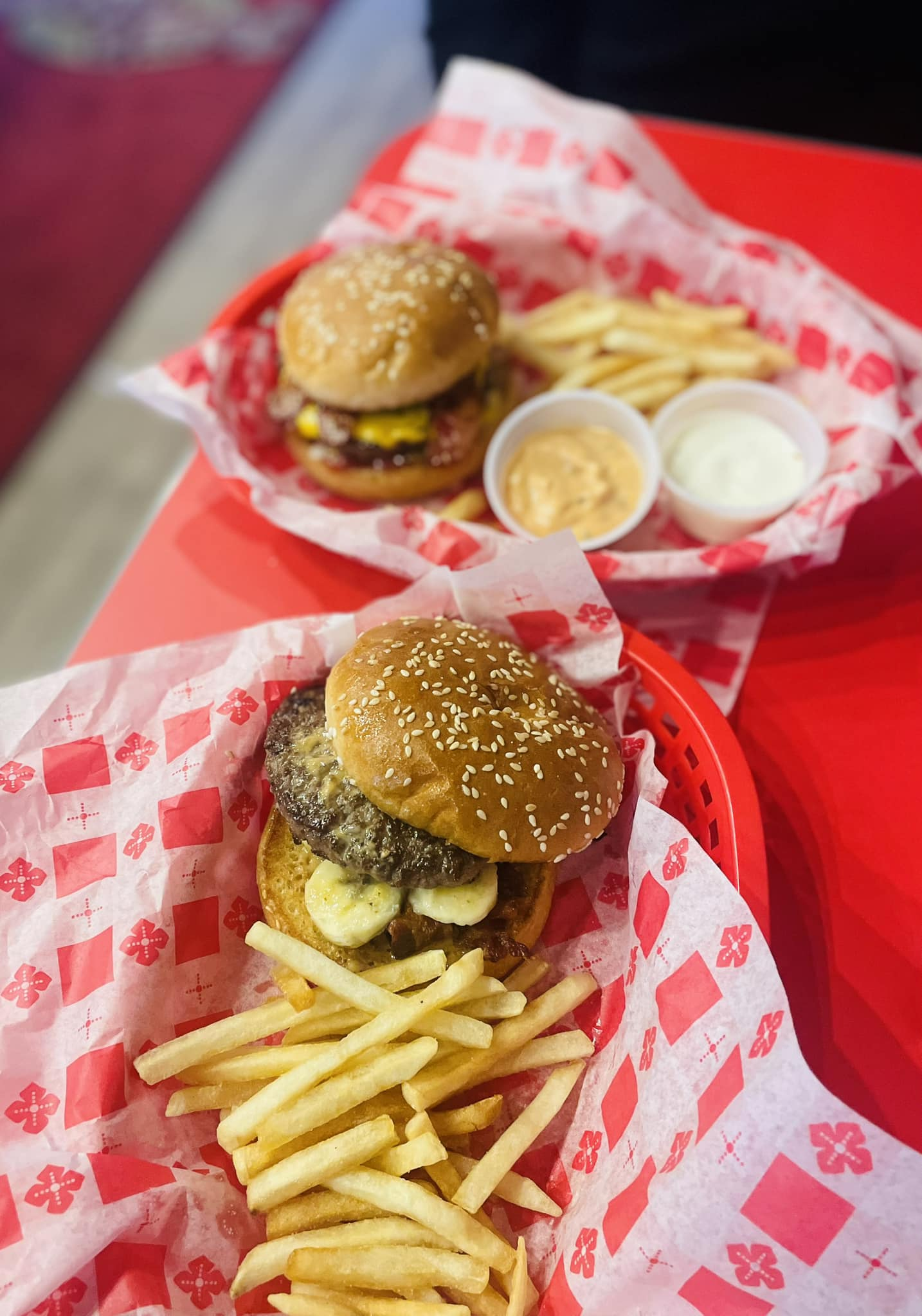 Two burgers with fries in red baskets, served with dipping sauces on a red table.