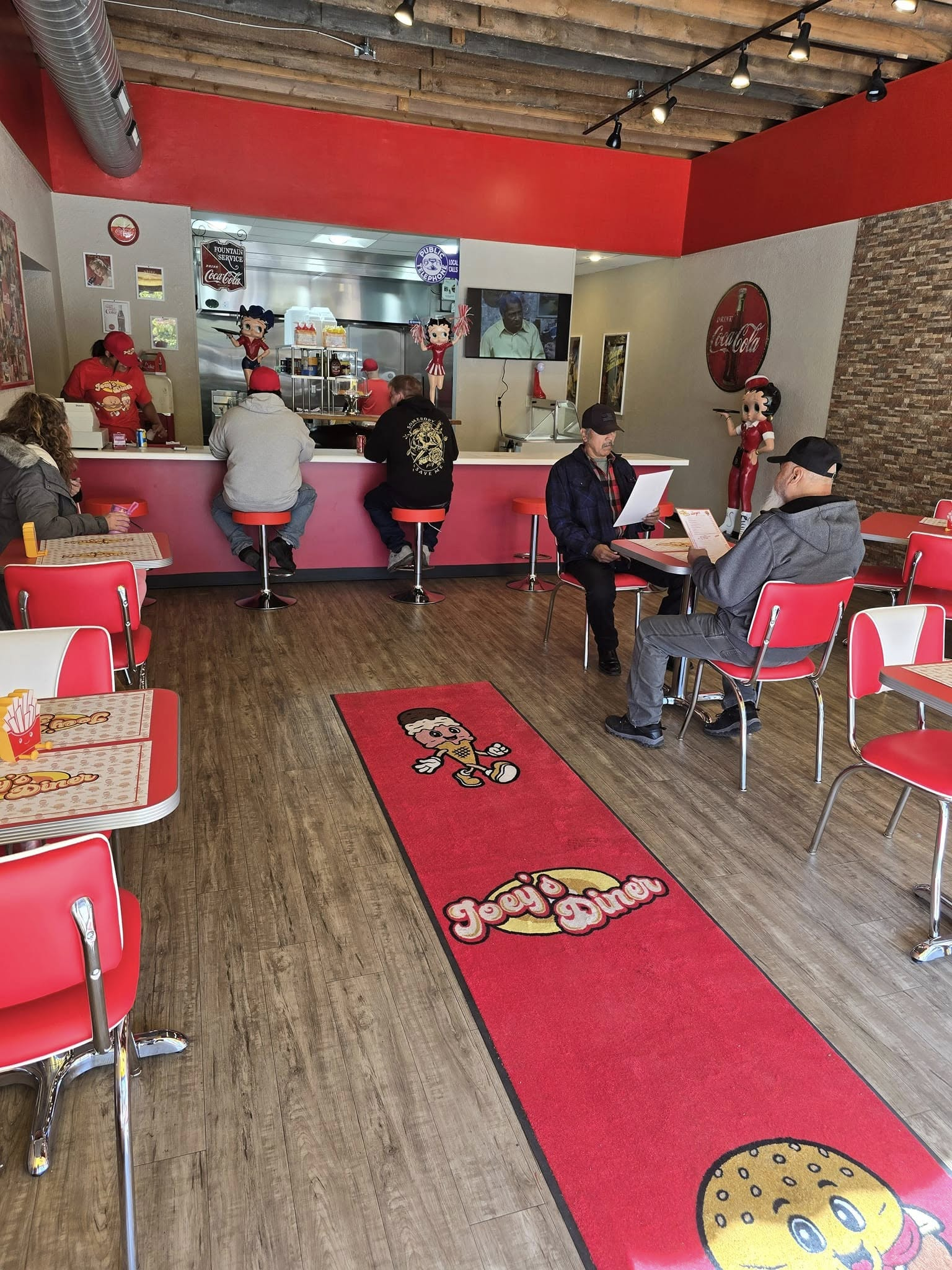 Interior of a retro diner with customers. Red decor, a rug, and counter seating are visible.