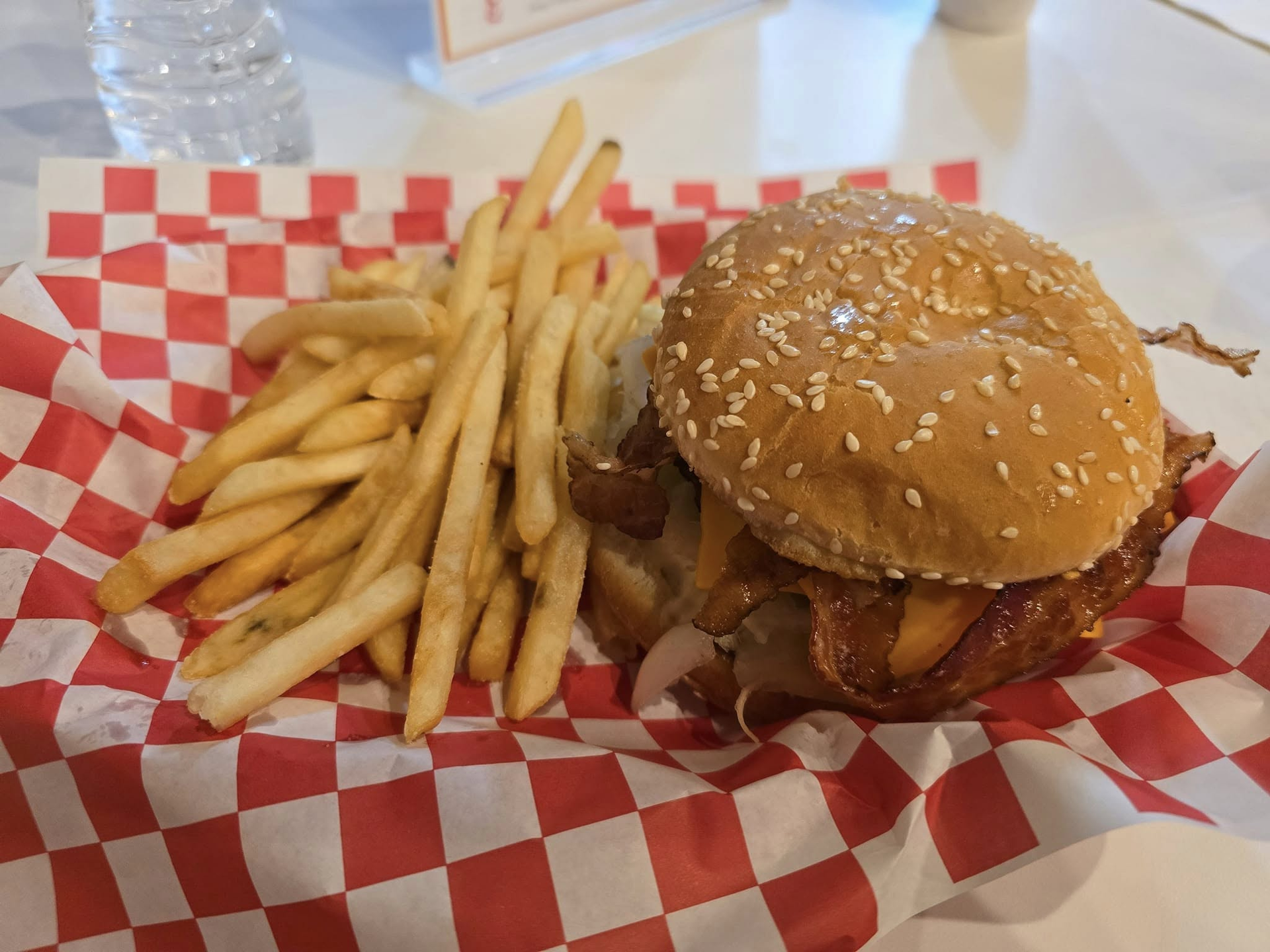 Burger with sesame seed bun, bacon, and fries in a red checkered basket.