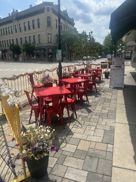 Red tables and chairs on a sidewalk patio in front of a building on a sunny day.