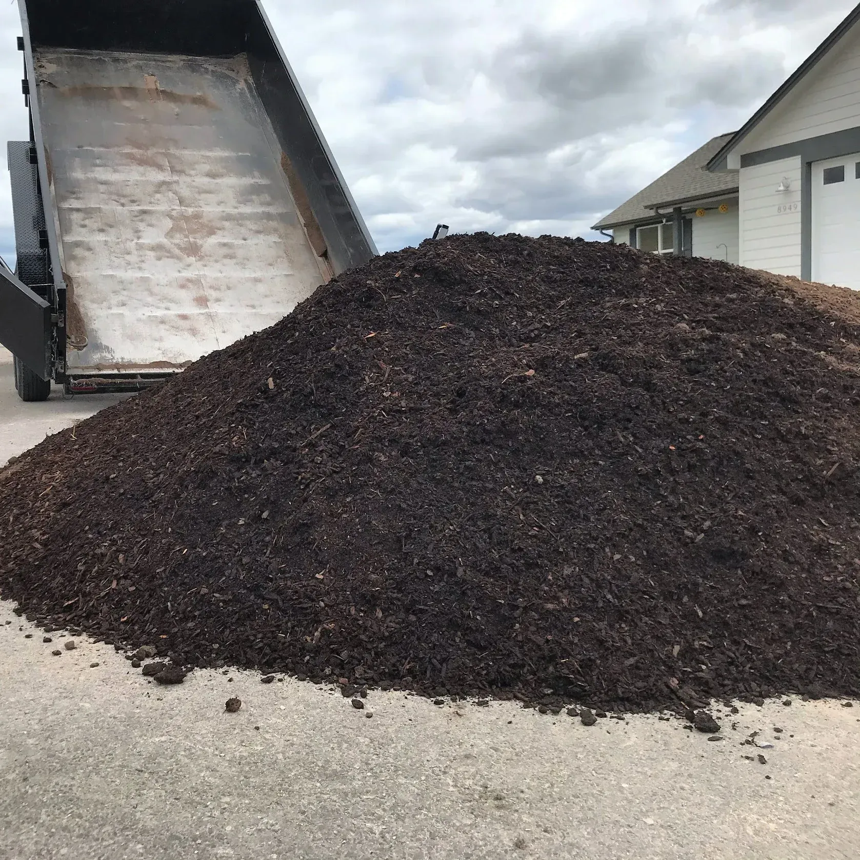 Dump truck unloading a large pile of dark brown mulch onto a concrete surface, near a house.