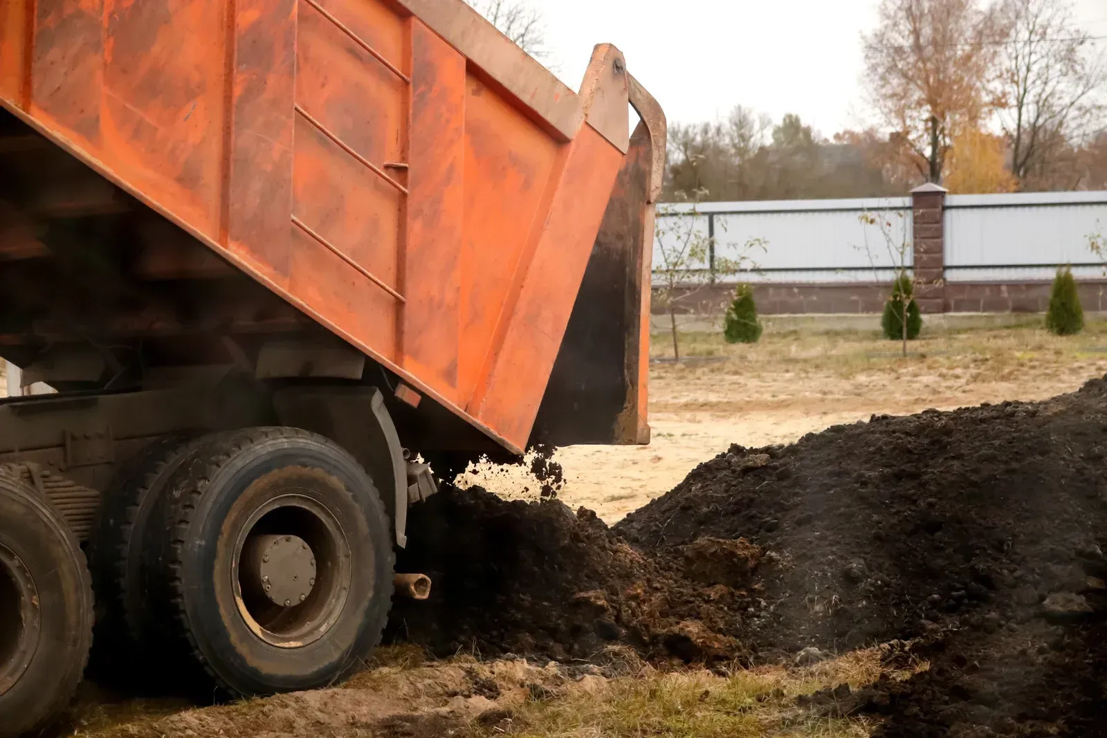 Dump truck unloading dark soil onto a dry, grassy area, near a fence.