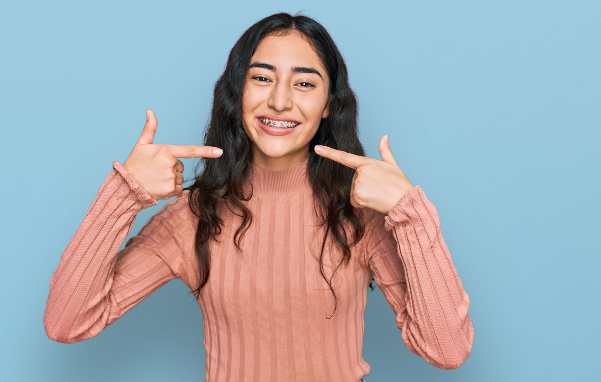 A girl with dental braces, wearing casual clothes, cheerfully showing and pointing at her mouth.