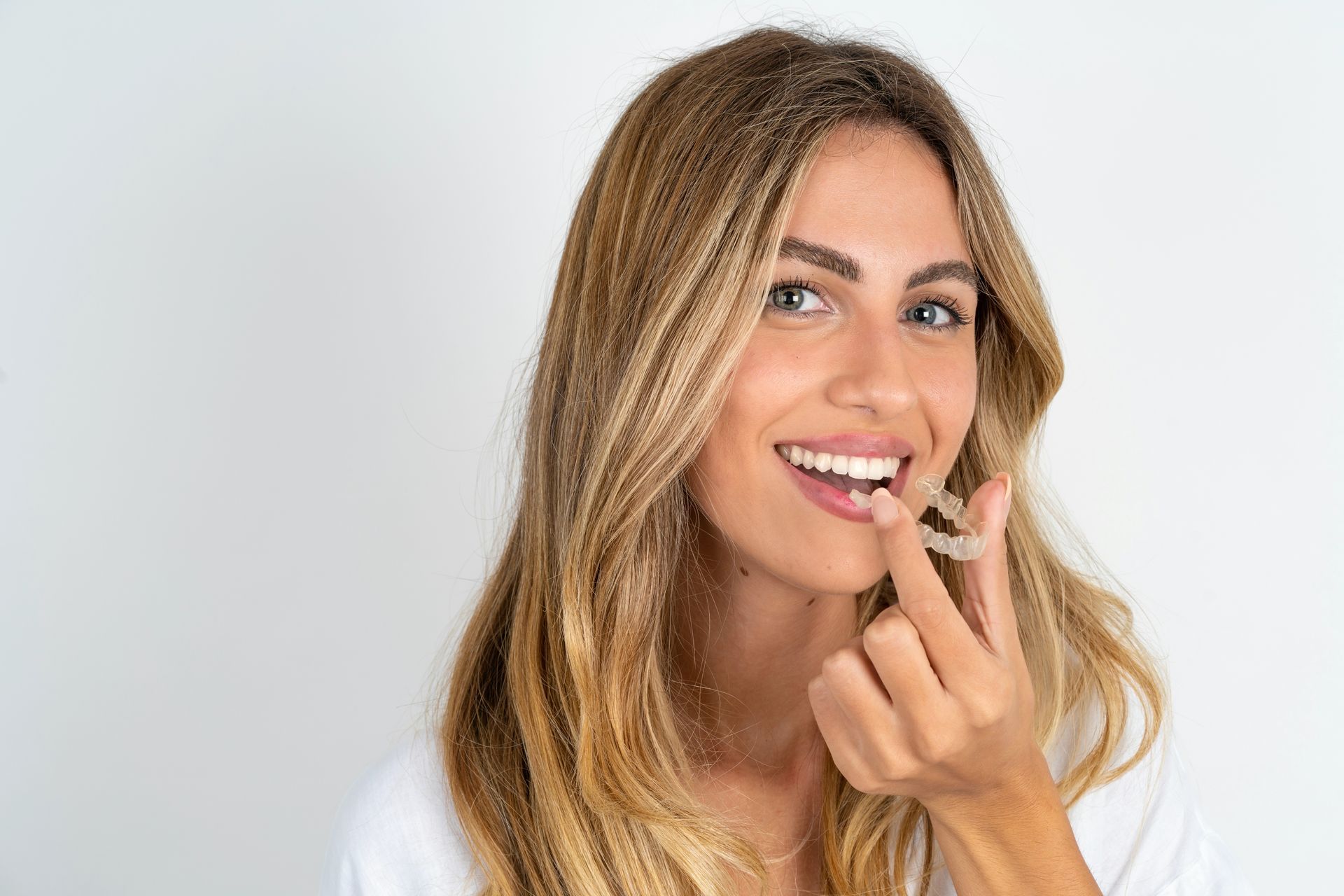 A smiling young blonde woman wearing a white shirt, holding an invisible aligner.