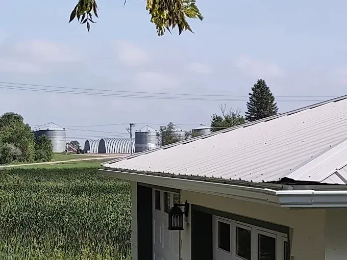White-roofed building overlooking a rural landscape with silos, power lines, and a field of crops under a blue sky.