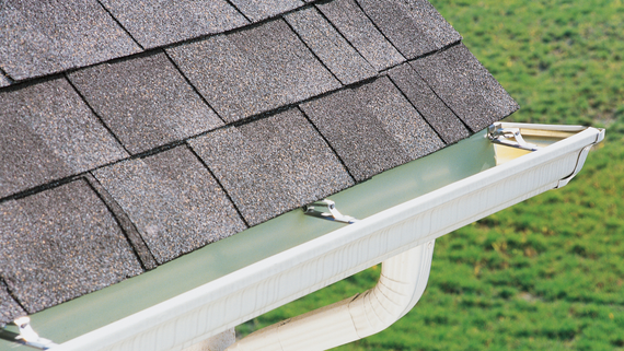 Close-up of a roof with gray shingles and a white gutter, downspout visible against a green field.