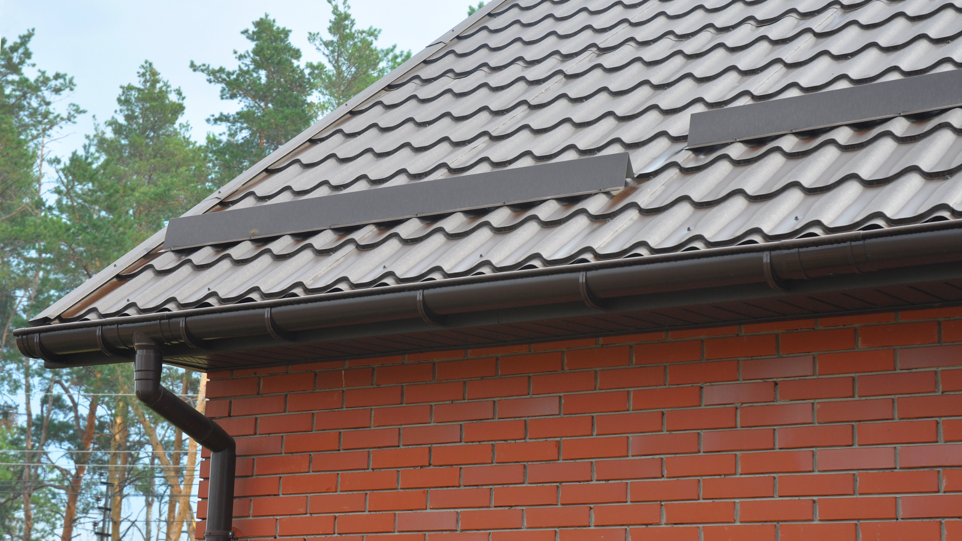 Brown roof tiles, gutters, and downspout on a brick building with trees in the background.
