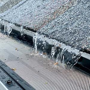 Water overflowing from a clogged roof gutter with a mesh guard, on a gray shingle roof.