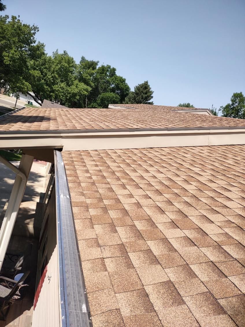 View of a brown shingle roof with a silver metal edge, under a blue sky, trees in the background.
