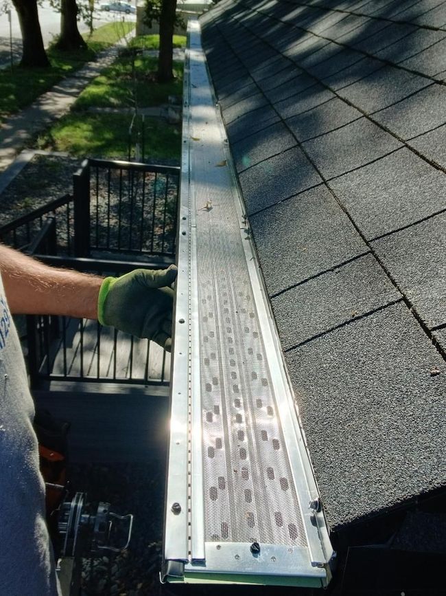 Person installing a metal gutter guard on a shingled roof, outdoors.
