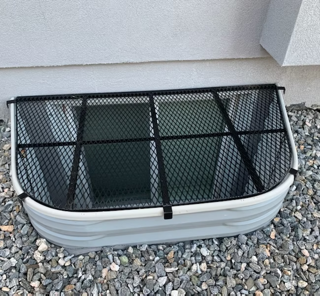 Window well with a black mesh cover, gray well, surrounded by gravel, against a white building.