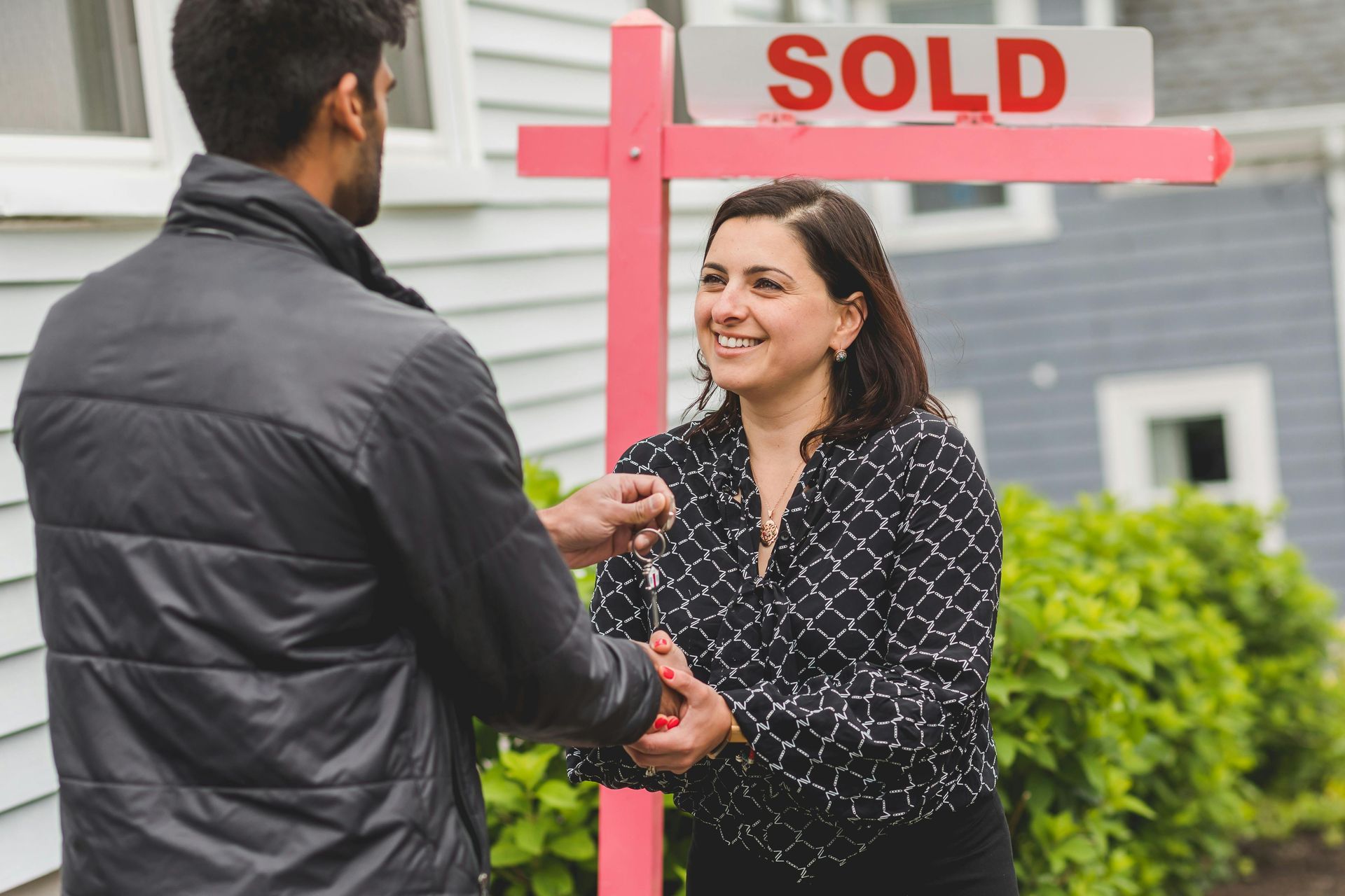 Two people shake hands beside a red SOLD real estate sign in front of a house.