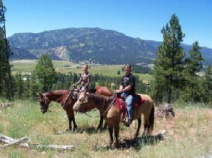 Two people are riding horses in a field with mountains in the background.